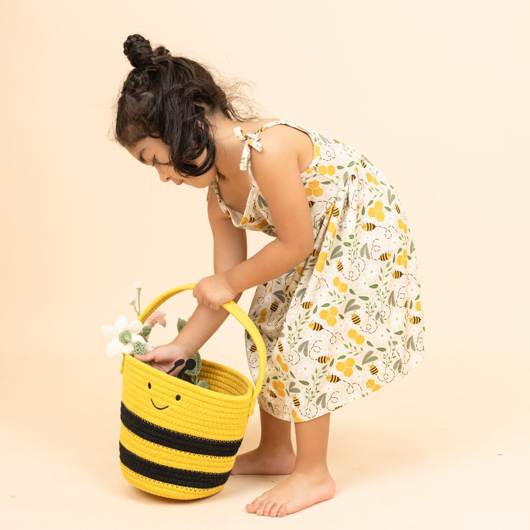 Young girl wearing a happy honey bee print bamboo sundress, reaching into a cute bee-themed basket filled with flowers. This lightweight and hypoallergenic dress is perfect for spring and summer adventures.