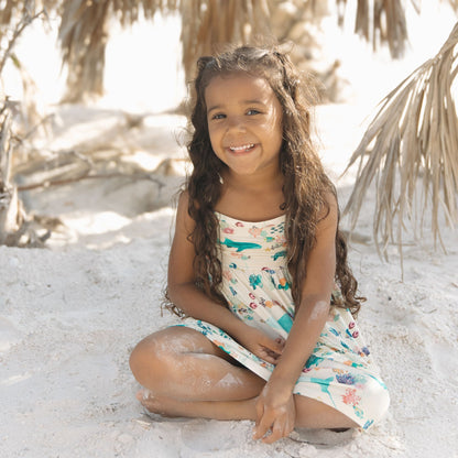 Toddler girl smiling while sitting at the beach in Emerson and Friends Coral Reef Bamboo Sundress, featuring a soft, breathable fabric with an ocean-themed print of sharks, sea turtles, and coral, designed for lightweight summer comfort and easy movement.