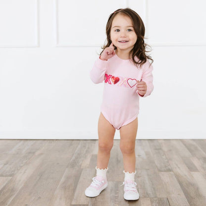 Child wearing a pink onesie with heart designs on a wooden floor.