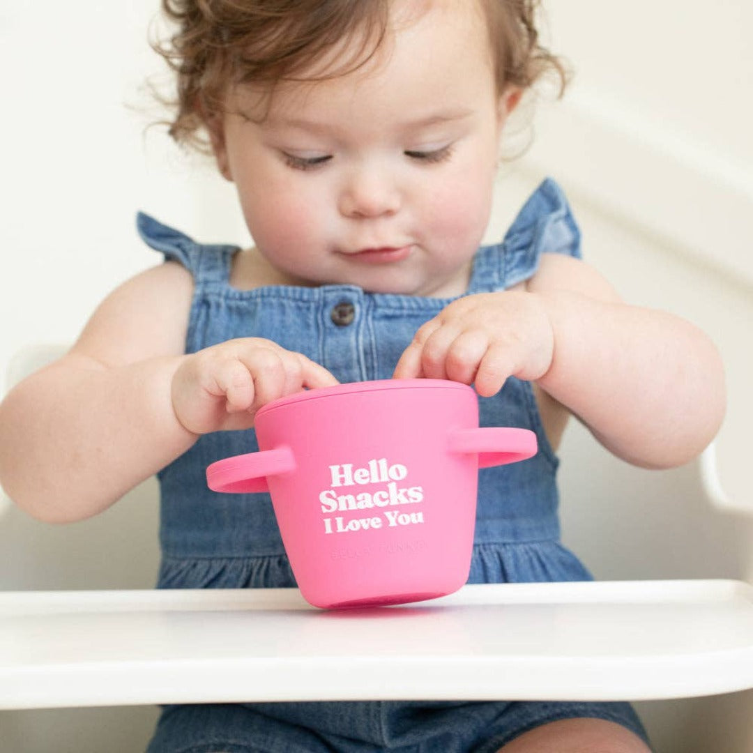 toddler in highchair grabbing snacks out of pink silicone snack cup that says hello snacks i love you