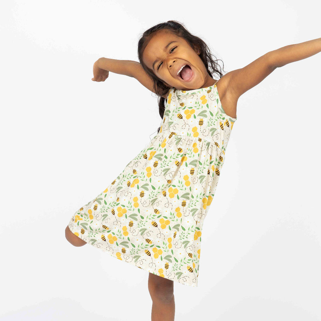 Excited young girl twirling in a lightweight bamboo sundress with a charming honey bee and honeycomb print. This breathable and hypoallergenic dress is perfect for warm-weather adventures.