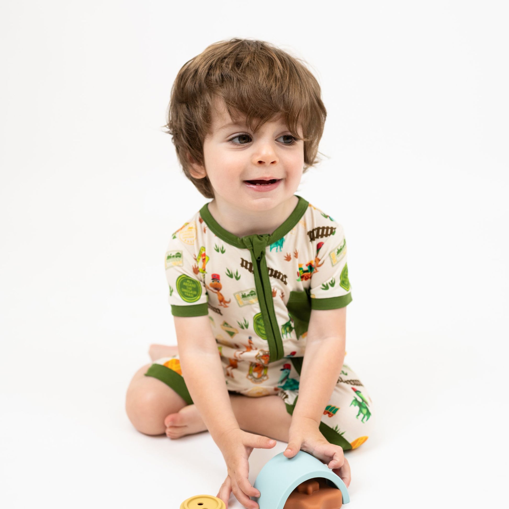 Toddler wearing our Emerson and Friends All Aboard the Dinosaur Train™ Bamboo Baby Shortie Romper featuring with green trim, and colorful orange, blue, and green dinosaurs on train tracks with "Dino Train" print.