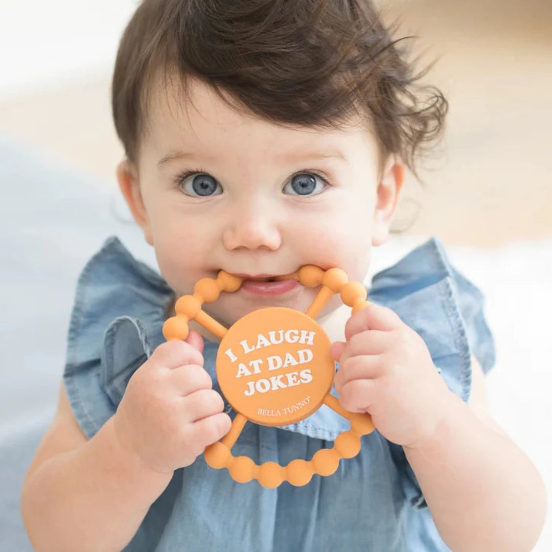 cute baby chewing on orange teething ring that says I laugh at dad jokes