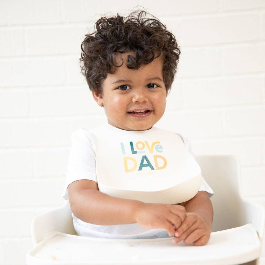 little boy sitting in highchair wearing a white silicone bib that reads I love dad in multi colors