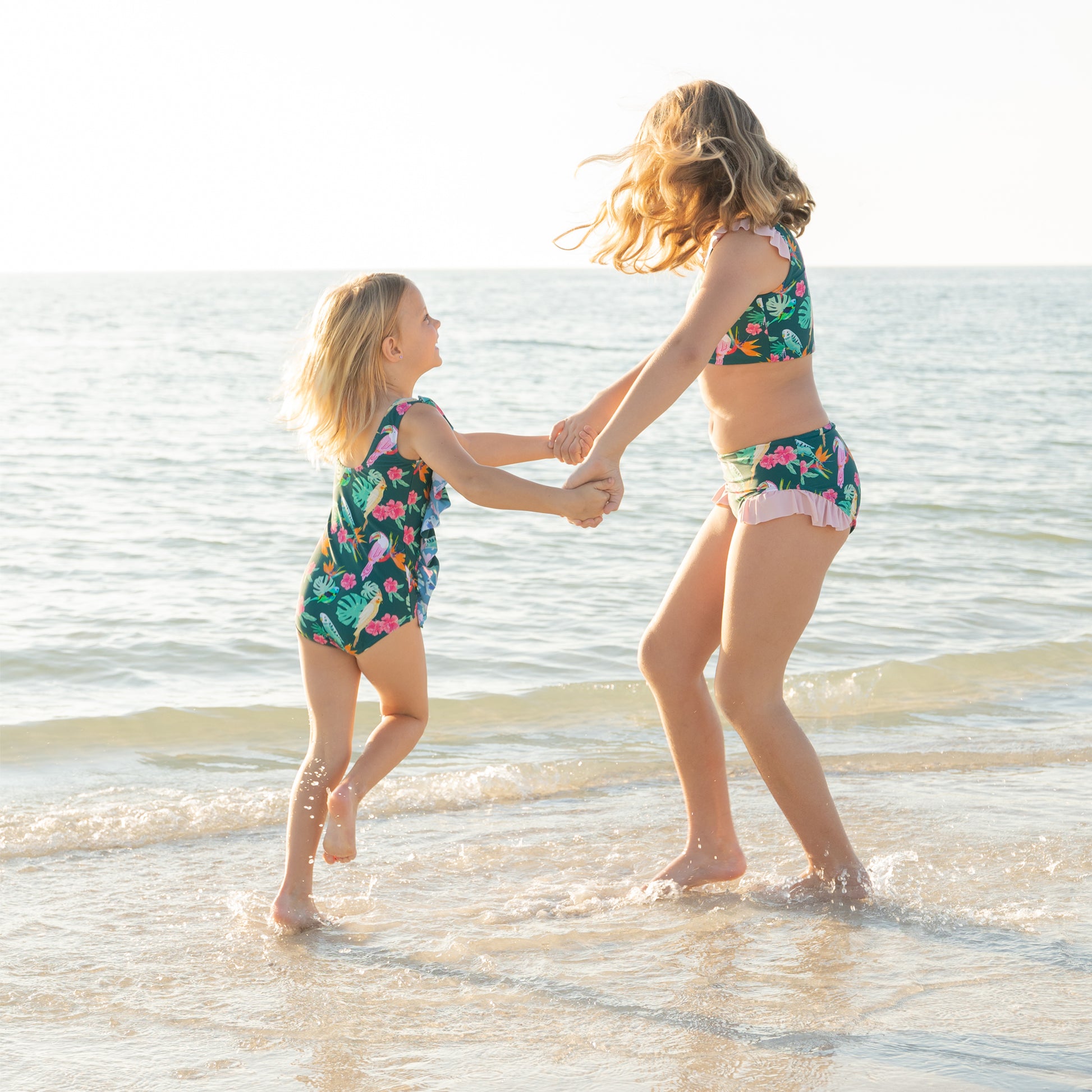 Two sisters playing on the beach in matching Isla Botánica tropical print swimwear, highlighting the ruffle one-piece and bikini styles.