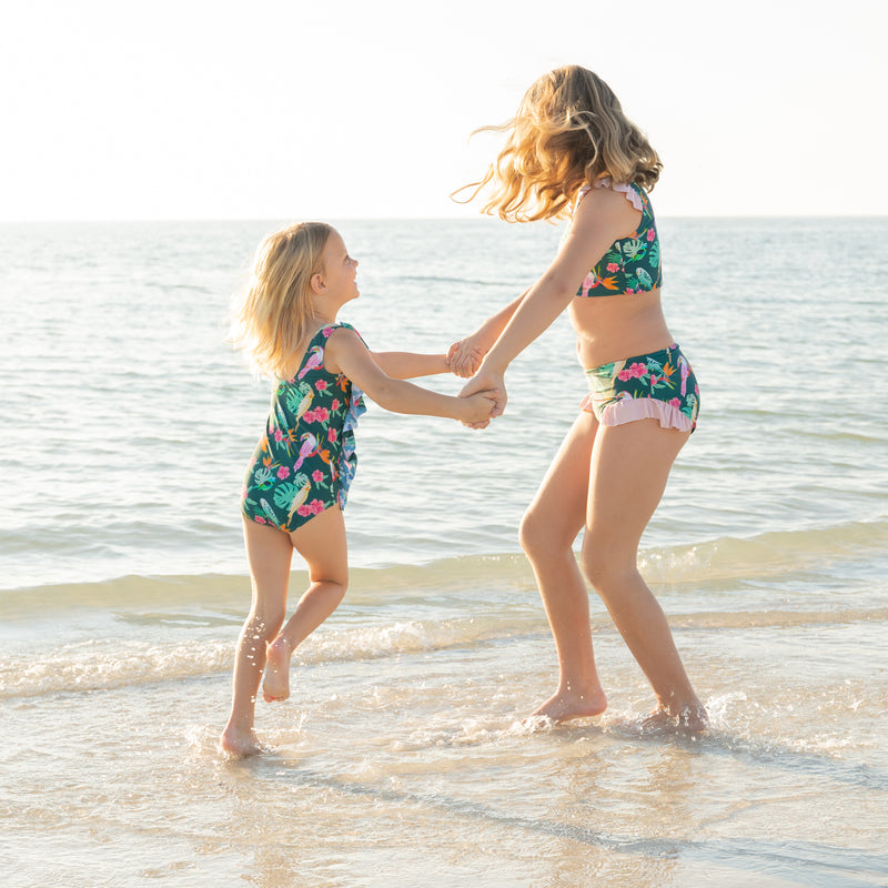 Two sisters playing on the beach in matching Isla Botánica tropical print swimwear, highlighting the ruffle one-piece and bikini styles.