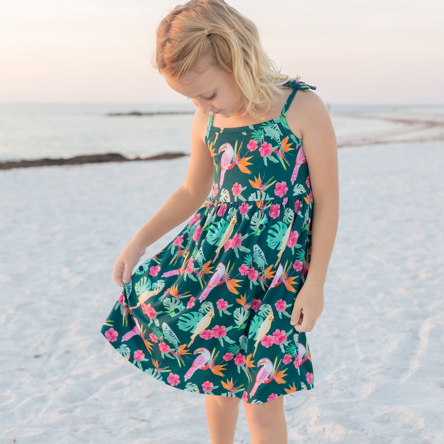 A young girl looking down at the flowy skirt of her Tropical Birds sundress, showing the play-ready and comfortable design.