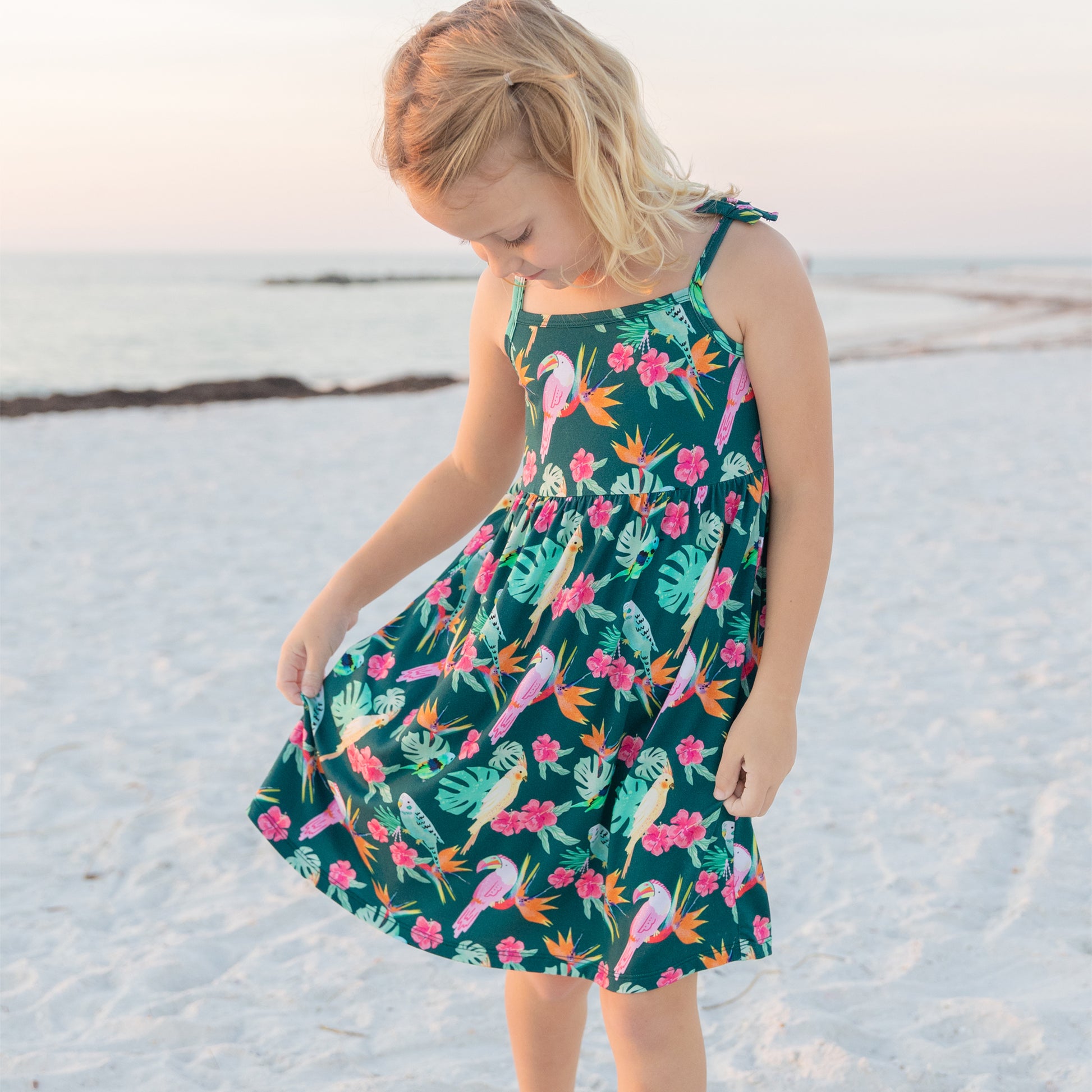 A young girl looking down at the flowy skirt of her Tropical Birds sundress, showing the play-ready and comfortable design.