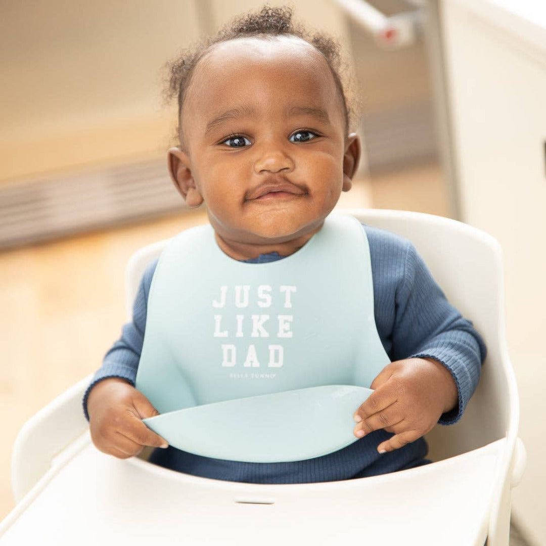 little boy in a highchair wearing a light blue silicone bib that says just like dad