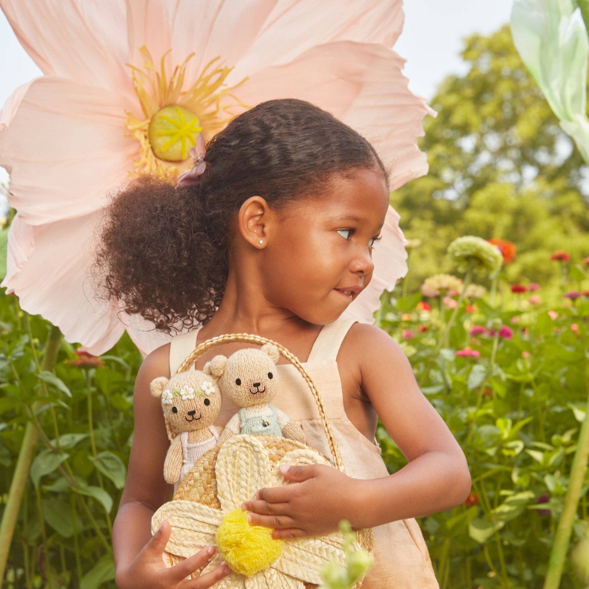 Young girl in a garden holding teddy bears and a basket, with a large flower in her hair.