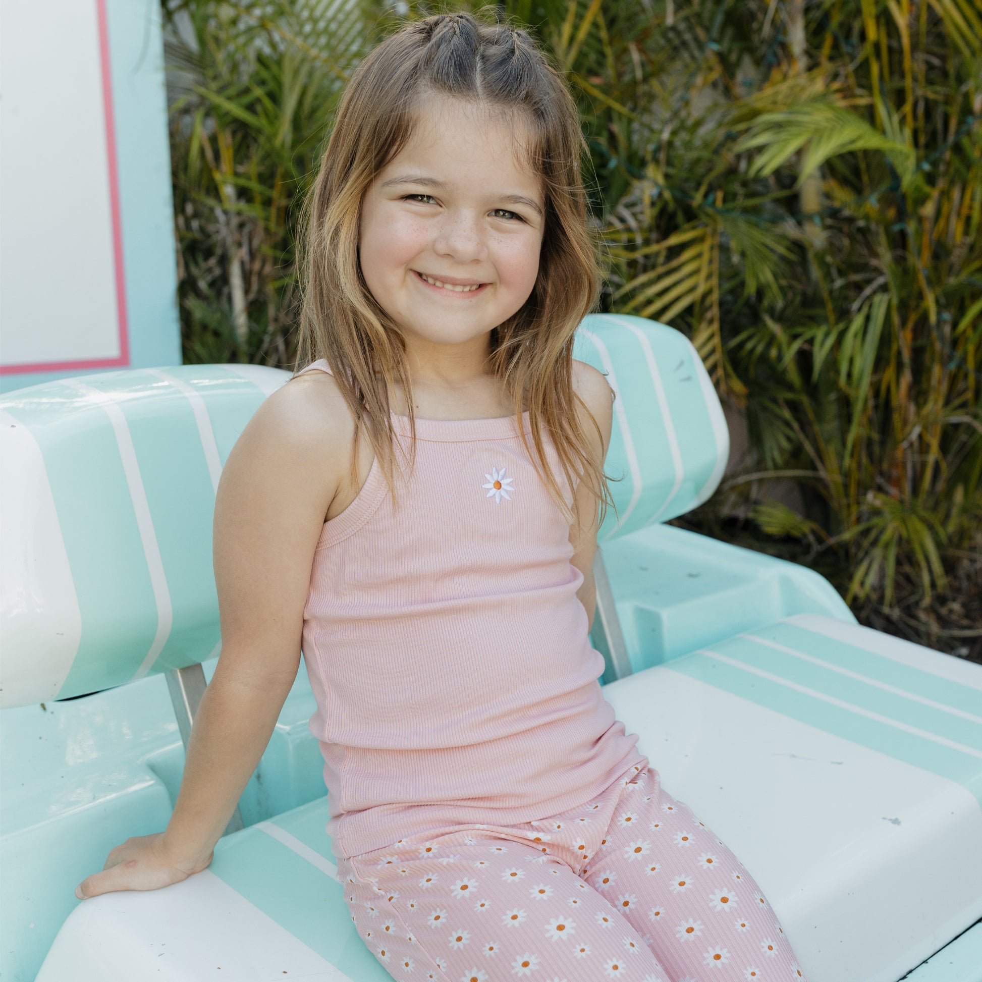Young girl in a pink dress sitting on a light blue chair with greenery in the background