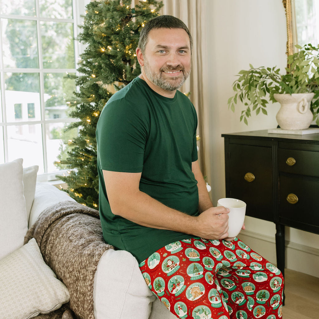 Man wearing Let It Snow Relaxed Bamboo Lounge Pajama Pants with a green shirt while sitting beside a Christmas tree.