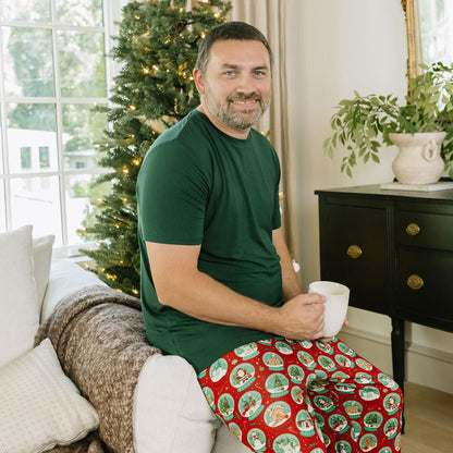Man wearing Let It Snow Relaxed Bamboo Lounge Pajama Pants with a green shirt while sitting beside a Christmas tree.