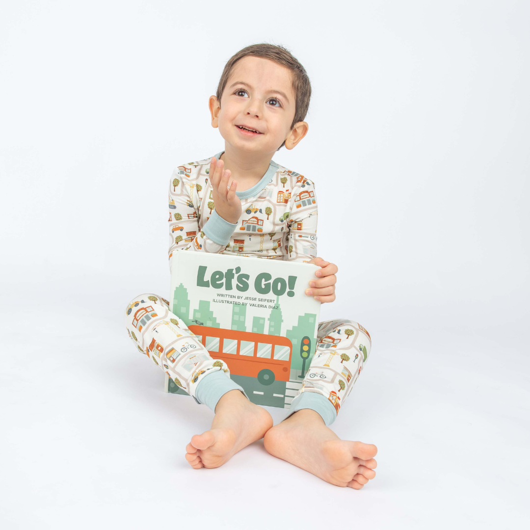 Kid holding out Let's Go Board Book in a white background and wearing our Let's go pajama bamboo set. 