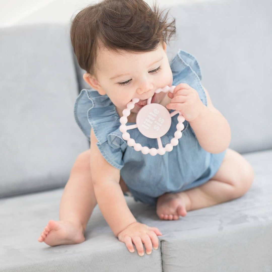 baby sitting on the couch chewing on a light pink round teether that says little sis