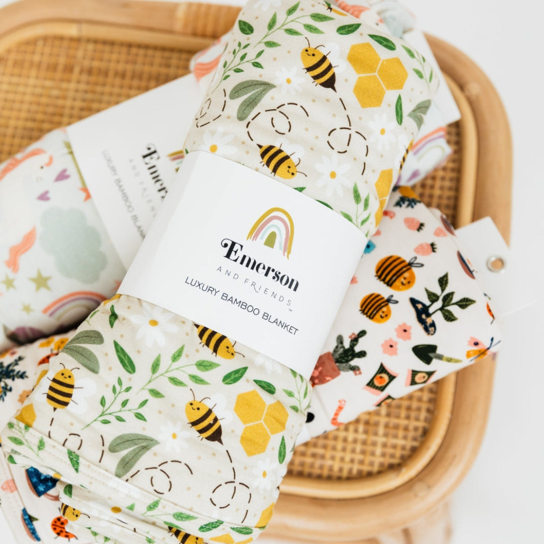 A beautifully rolled Happy Honey Bees luxury bamboo blanket by Emerson and Friends, featuring a charming bee and honeycomb design, placed on a rattan tray alongside other soft and breathable bamboo fabric items