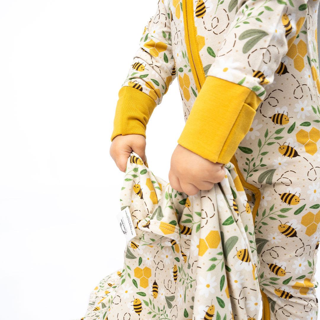 A close-up of a baby holding a soft, hypoallergenic bamboo blanket in a Happy Honey Bees print, featuring adorable bees, honeycombs, and floral accents. The cozy fabric and fold-over cuffs add extra comfort.