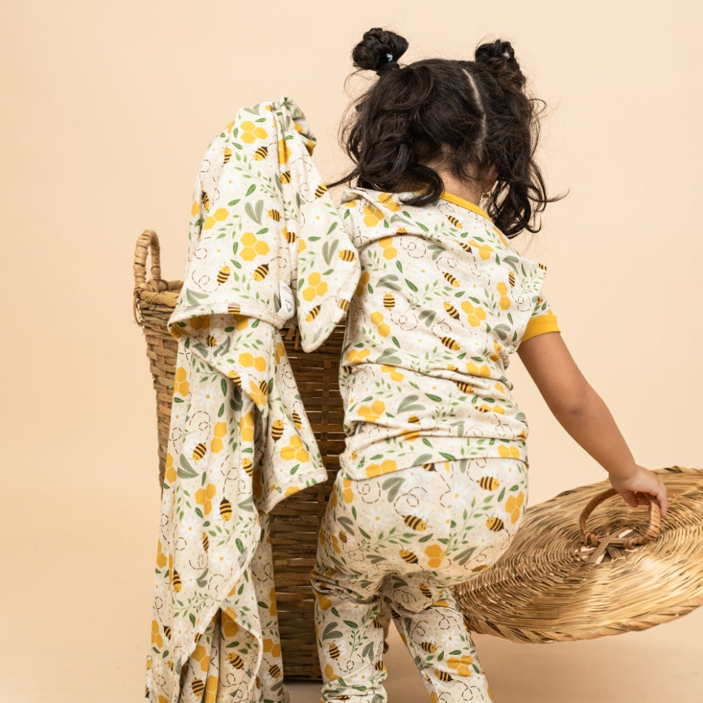 A toddler wearing a matching Happy Honey Bees bamboo pajama set, reaching into a woven basket while holding a soft, breathable luxury bamboo blanket with an adorable bee and honeycomb print.