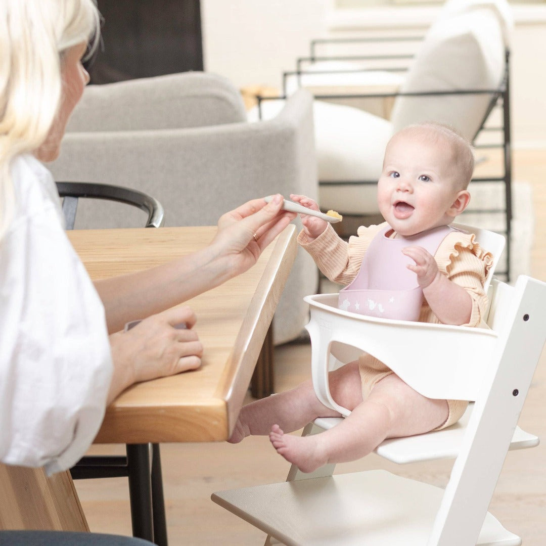 baby girl in a highchair being fed by her mom while wearing a light purple silicone bib with unicorns on it