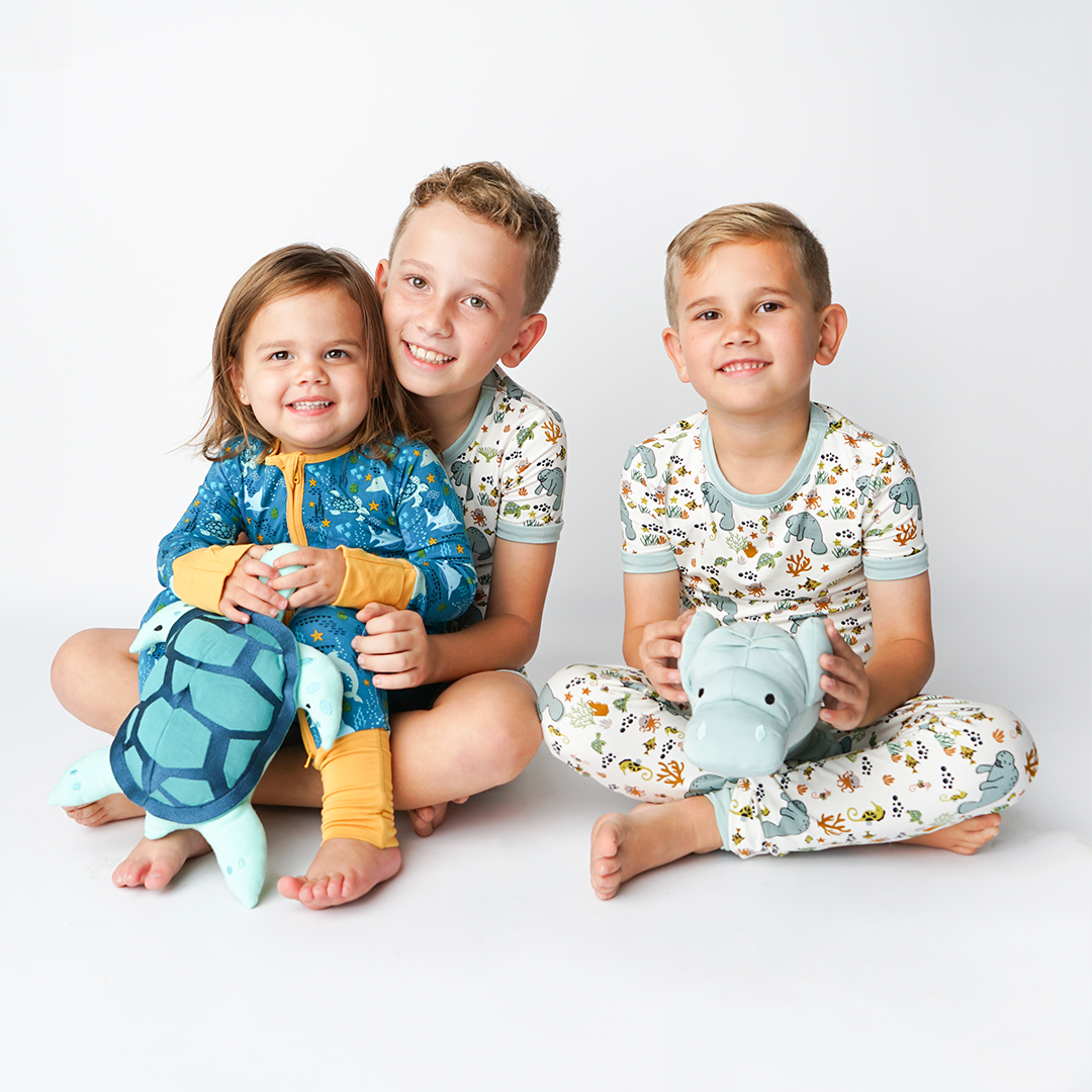 3 kids sit on the floor and smile for the camera. a little girl sits in the lap of an older boy. they both smile and the girl wears the "ocean friends" convertible and holds Toby the sea turtle. the boy next to them wears the "manatee" matching pajama set and holds Max the manatee. 