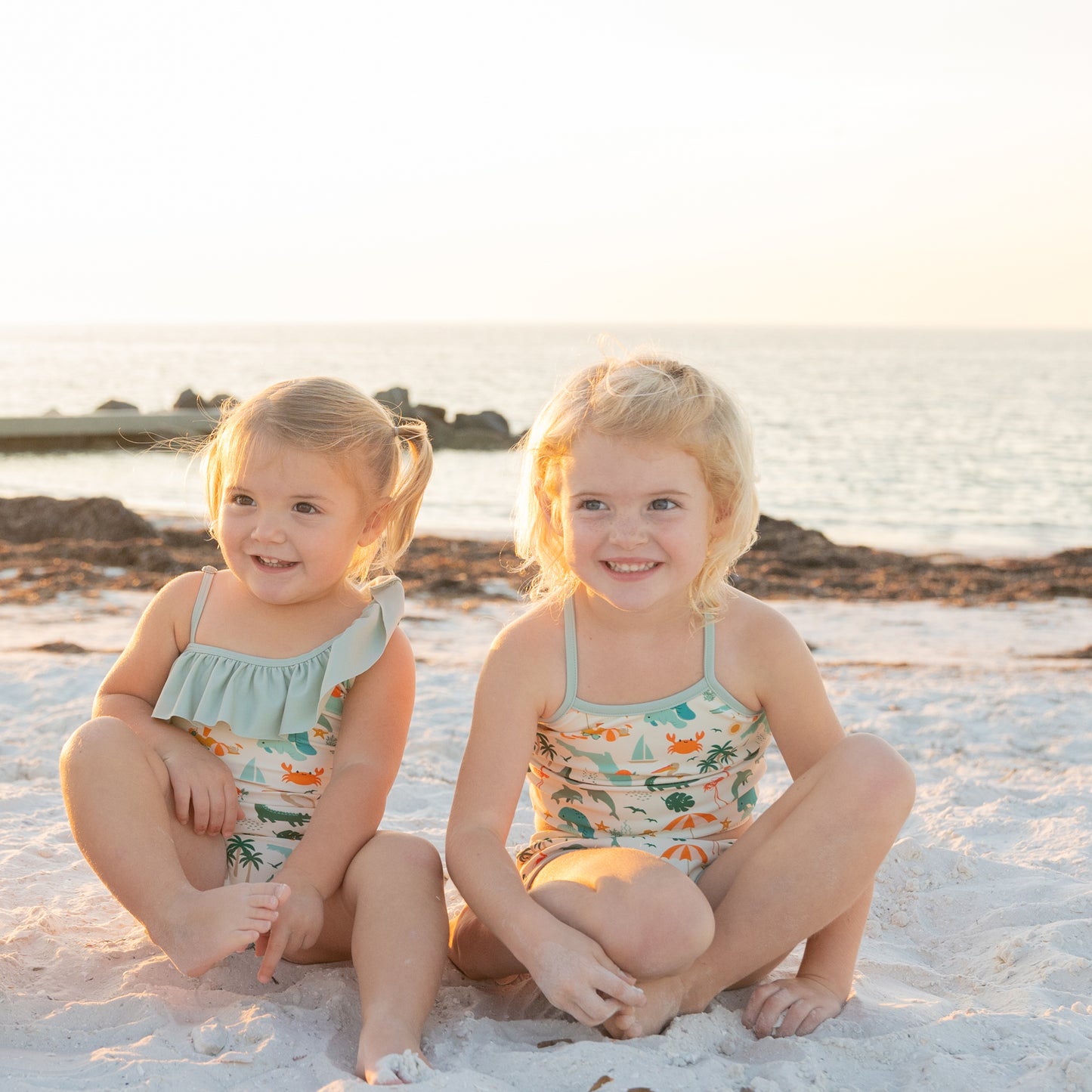 Two young girls sitting on the sand in coordinated Explore Florida swimwear, showing the athletic and comfortable fit.