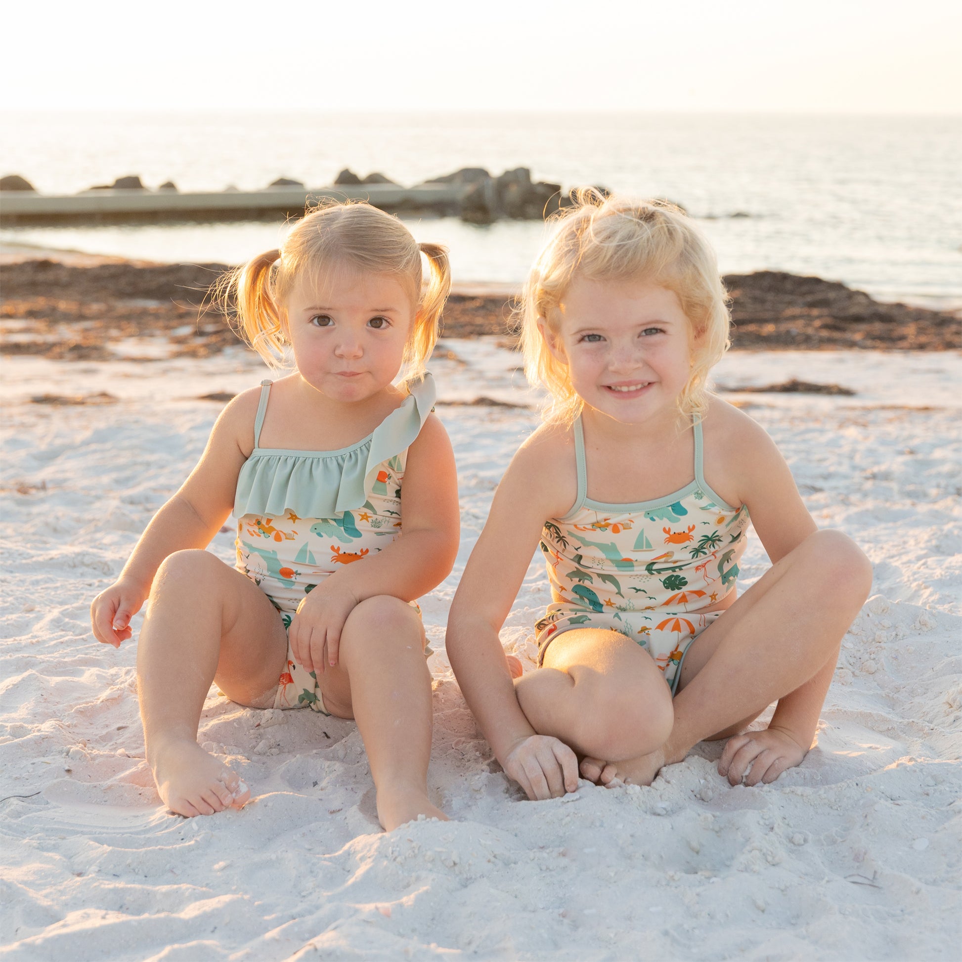 Two young girls sitting on the beach in matching Explore Florida swimsuits, perfect for coordinated sister beach photos.