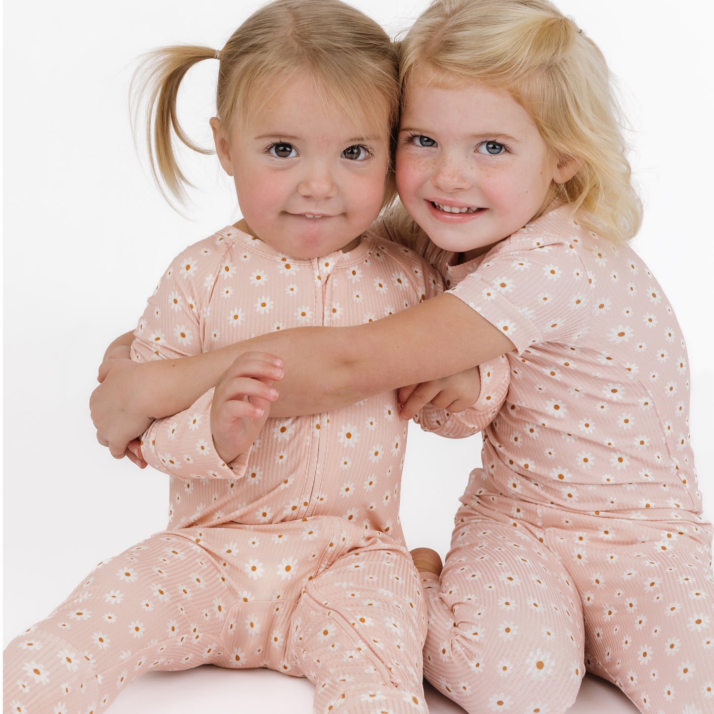 Two young girls wearing matching pink outfits with a subtle pattern, sitting close together on a white background.
