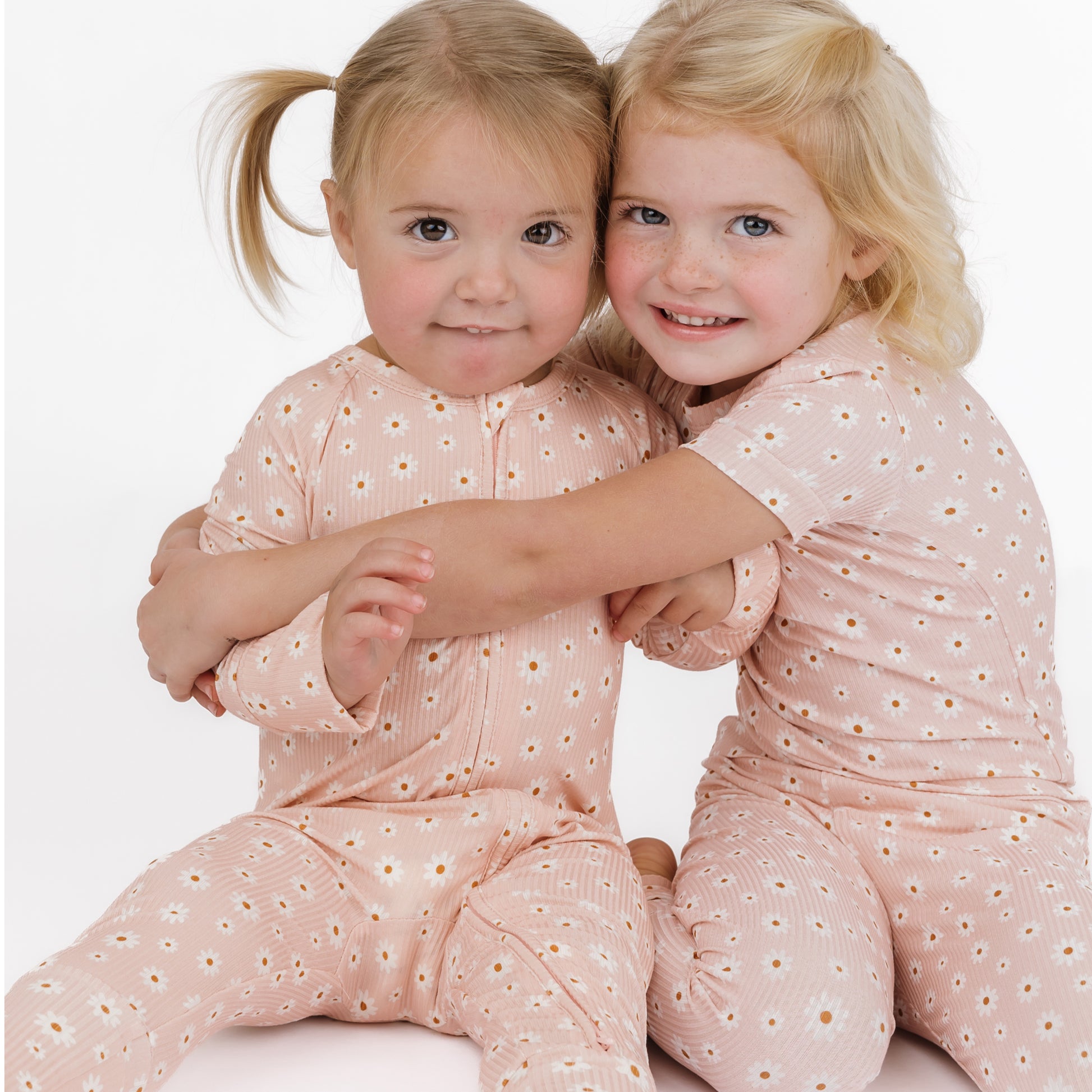Two young girls wearing matching pink outfits with a subtle pattern, sitting close together on a white background.