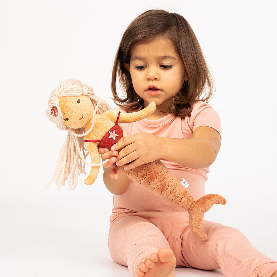 girl plays with Adriana the stuffed plush mermaid. she is a blonde mermaid with a red top and orange tail. 