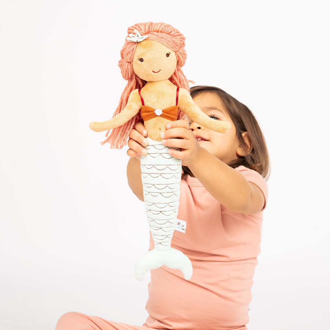 young girl holds up Cordelia the mermaid.  Cordelia is a red-head mermaid with an orange top and blue tail. 
