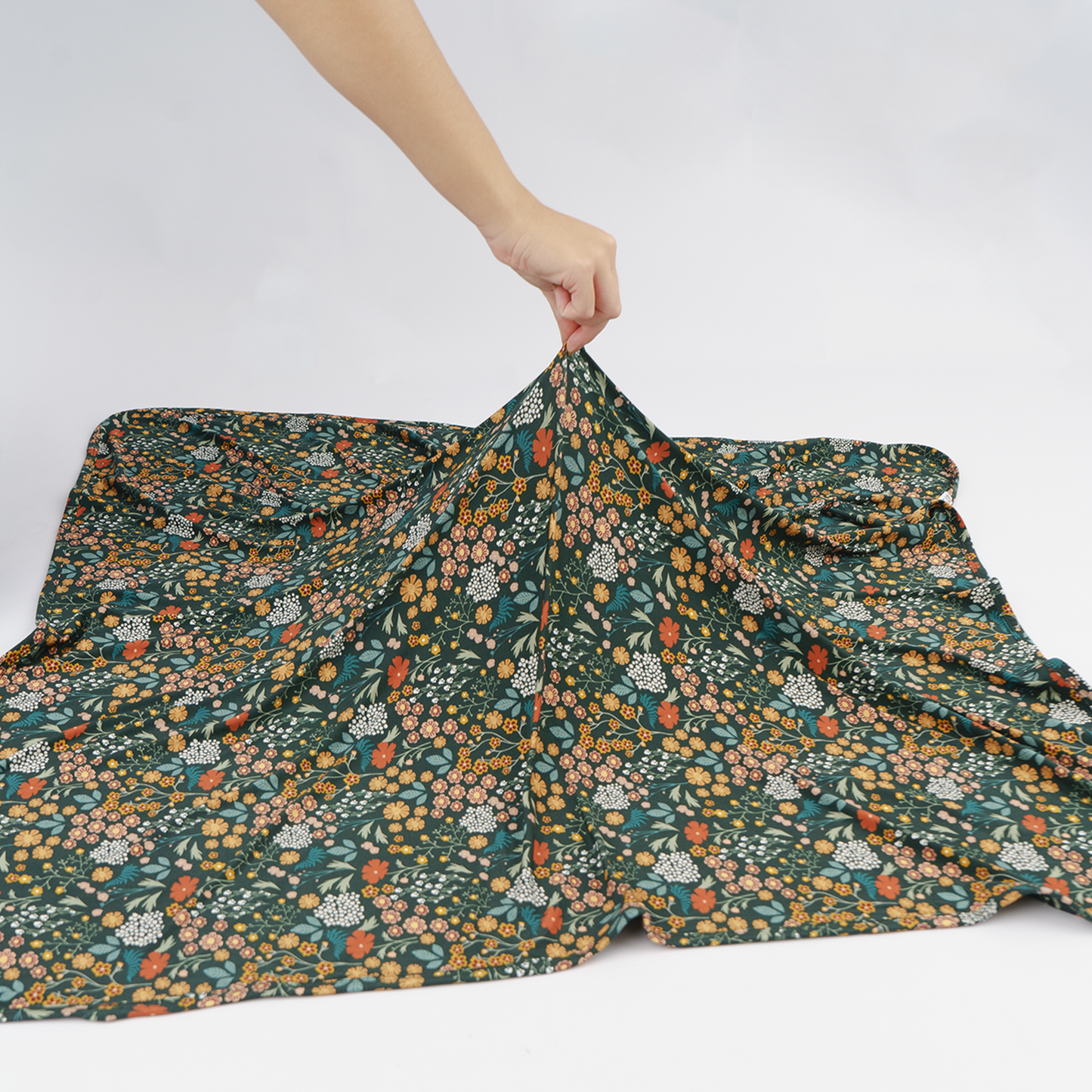 A dark green floral blanket is spread out on a white studio floor with a person's hand holding the top corner to show its drape and pattern.
