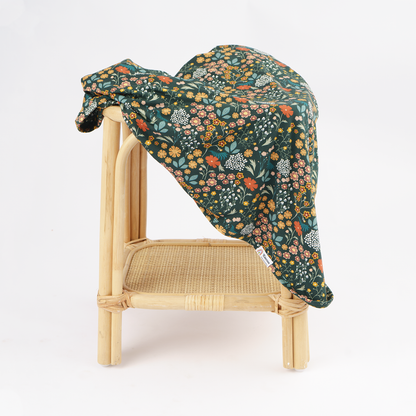 A dark green floral blanket draped over a light natural wood rattan chair against a clean white background.