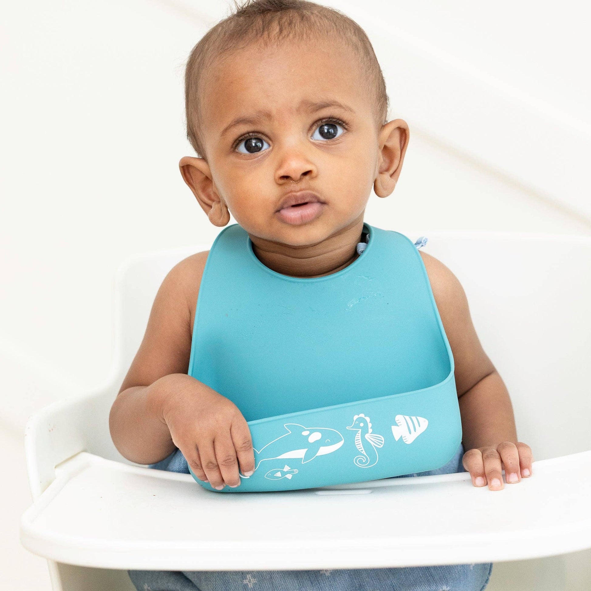 little boy sitting in his highchair wearing a blue silicone bib with white sea animals along the bottom edge