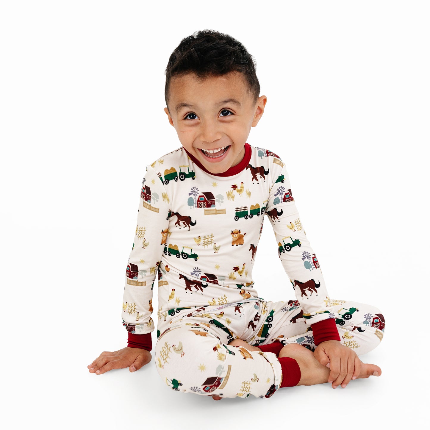 Child sitting in On the Farm bamboo long sleeve pajama pants set with barnyard print of horses, tractors, and cows.