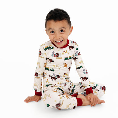 Child sitting in On the Farm bamboo long sleeve pajama pants set with barnyard print of horses, tractors, and cows.