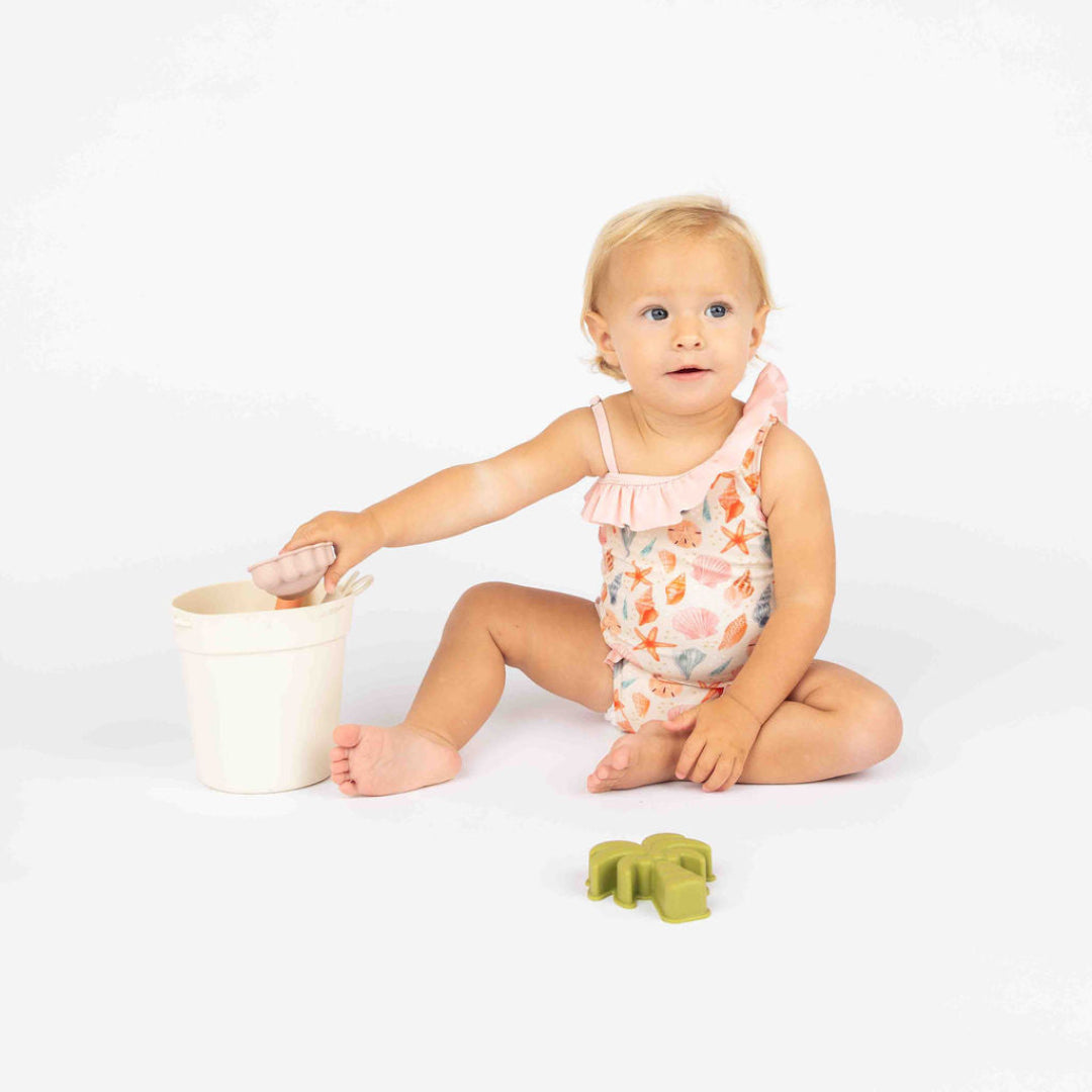 Baby girl sitting down on a white background wearing our sandy Seashell One piece swim suit with ruffles on the light pink truffles. 