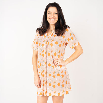 a brunette poses with one hand on her hip. she is wearing the "freshly squeezed" nightgown. the "freshly squeezed" print has an assortment of full and half cut oranges scattered around. there is also flower heads and flower stems that intermingle within the print. this is all space out around a pink background space. 