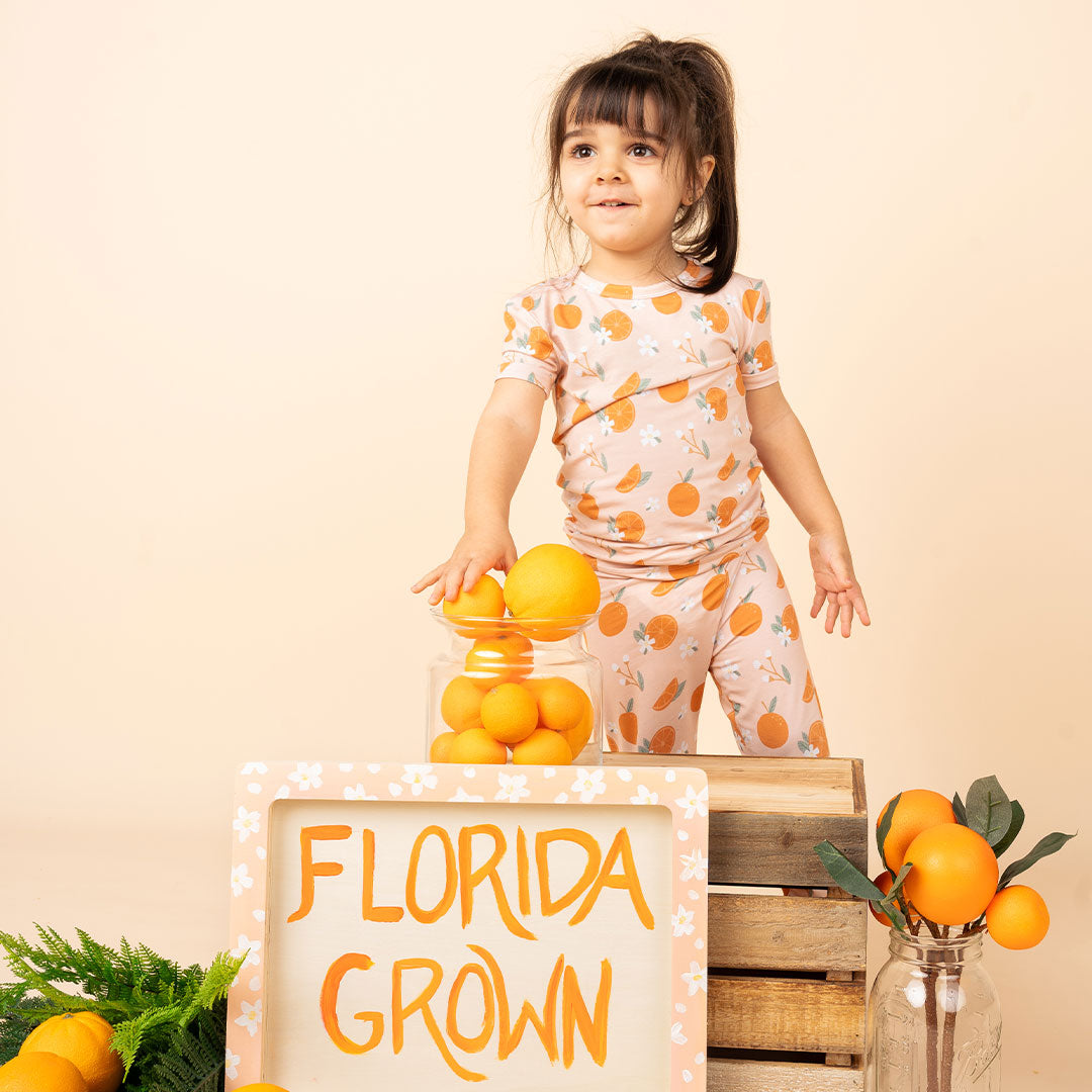 a young toddler girl stands on top of a wooden crate, resting her hand on a jar of oranges. she is wearing the "freshly squeezed" 2-piece matching set. there are oranges and leaves scattered around her and a sign that reads - Florida grown. 
