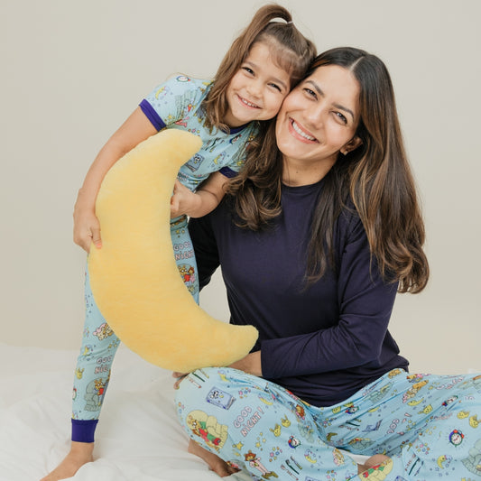 A heartwarming moment captures the mother with daughter smiling, with them wearing our PBS Good Night Arthur™ collaboration featuring pajamas adorned with Arthur and friends in a cozy bedtime scene complete with storybooks, toothbrushes, bunny slippers, a sleeping puppy, crescent moons with sleep caps, twinkling stars, and the words "GOOD NIGHT!" on a soft blue background.