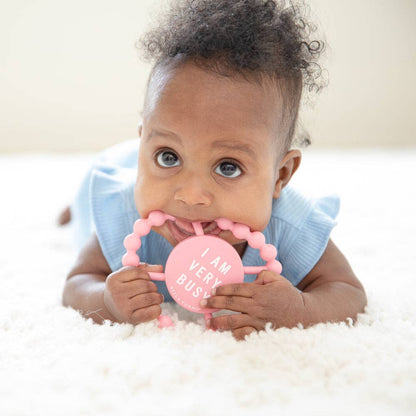 Baby holding a pink teething ring with text, lying on a white surface.