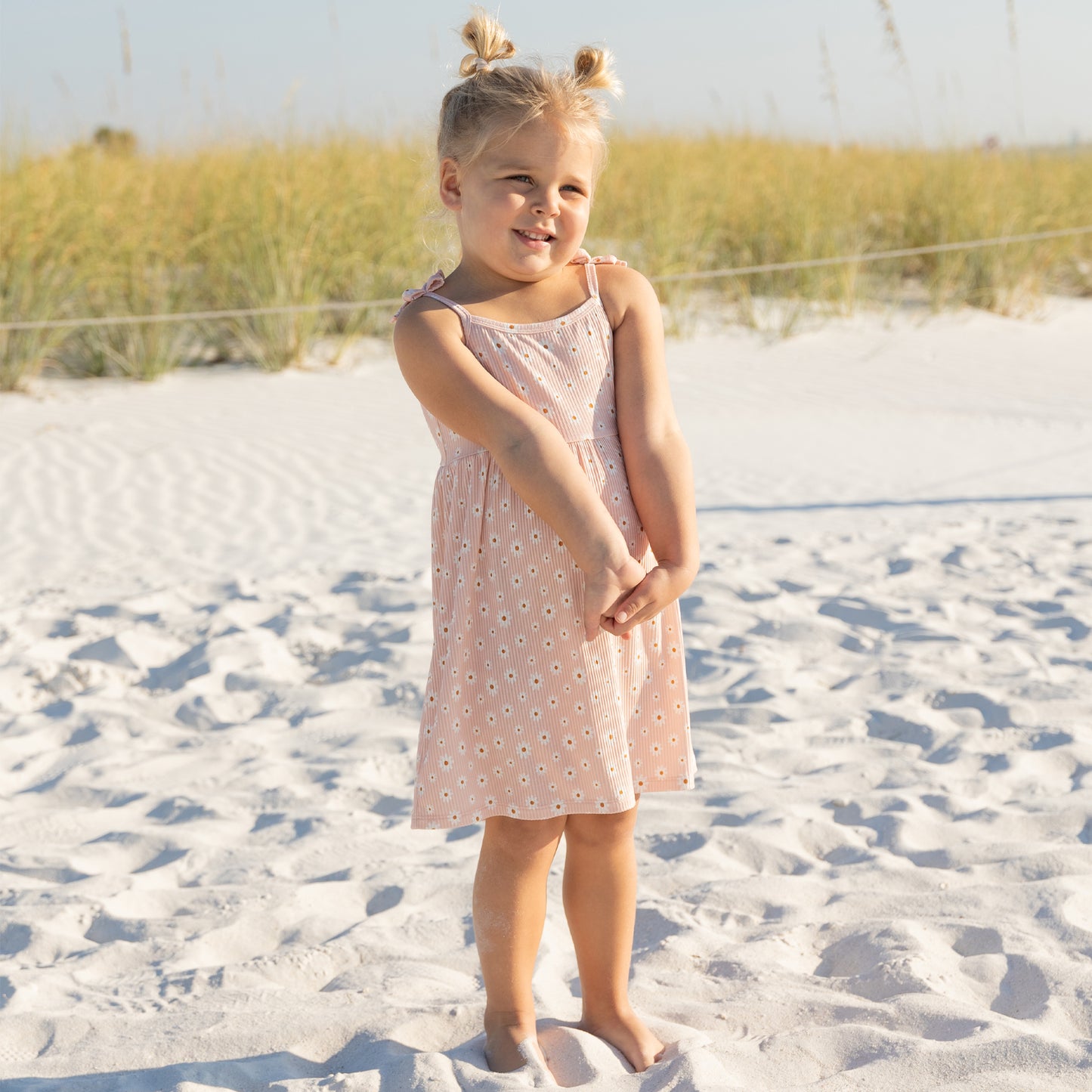 Young girl in a pink dress standing on a sandy beach with grass in the background