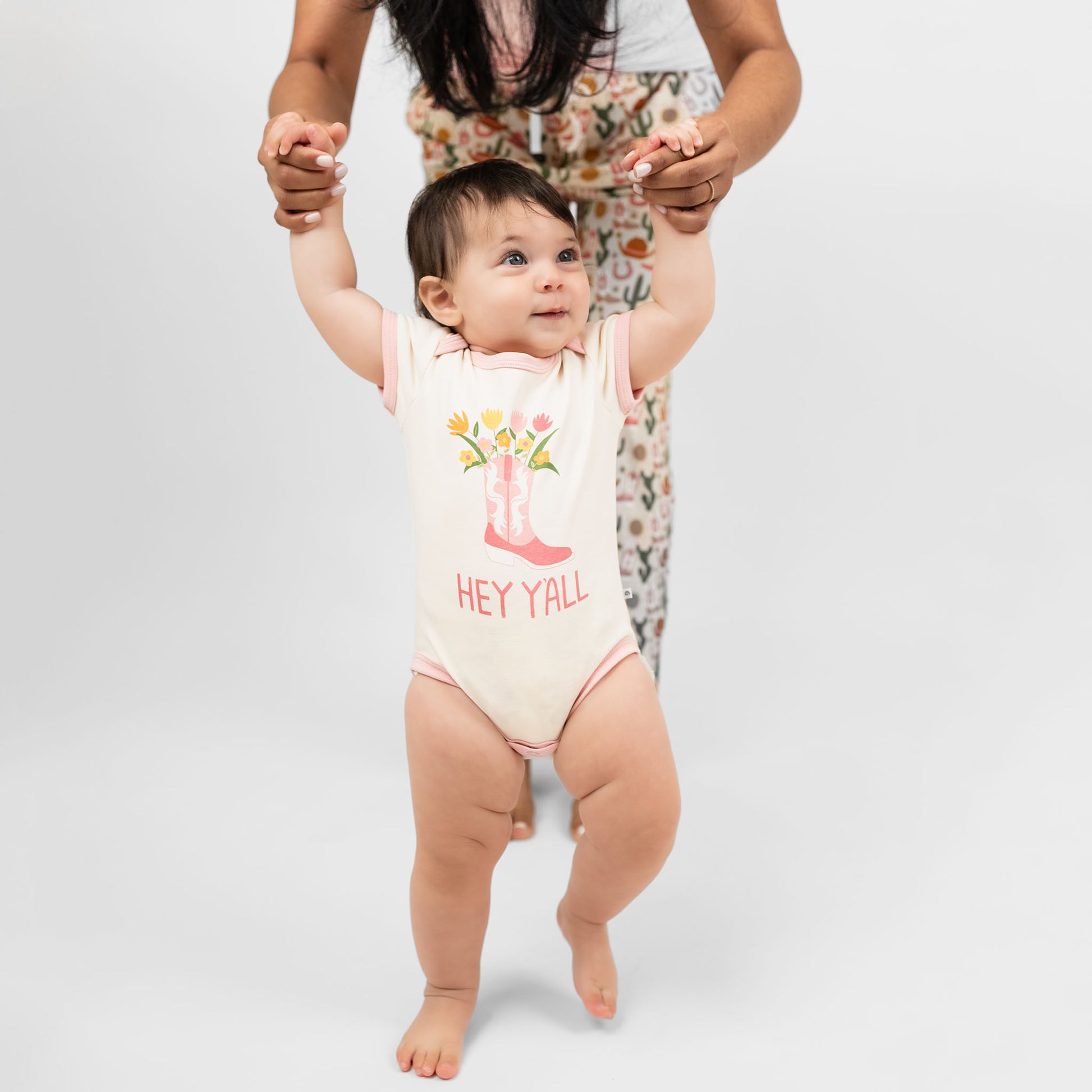 Cute baby girl wearing our Hey Y'all onesie paired with a pink cowgirl boots print and floral accents, capturing the essence of rodeo and western style for a perfect outfit.