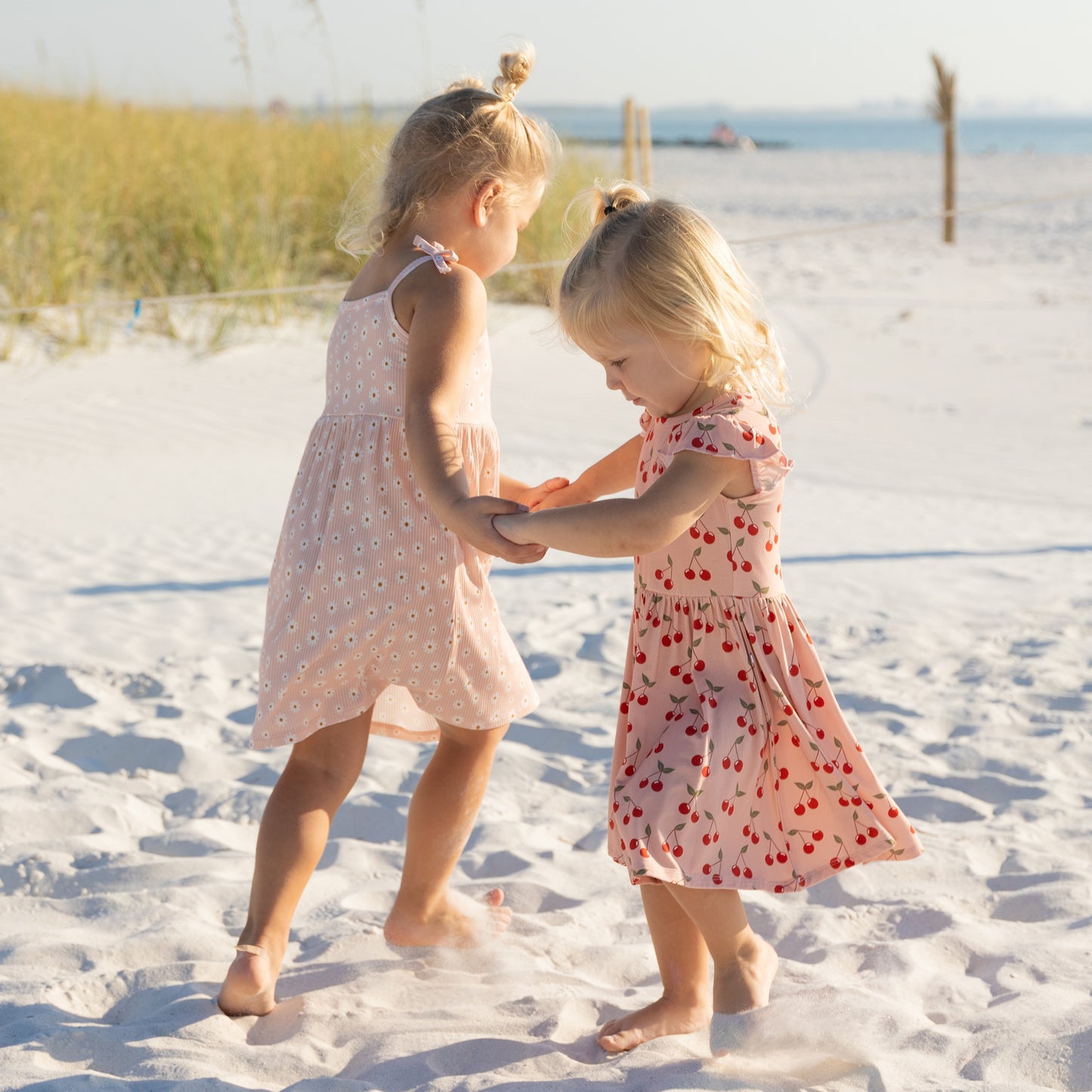 Two young girls in floral dresses standing on a sandy beach.