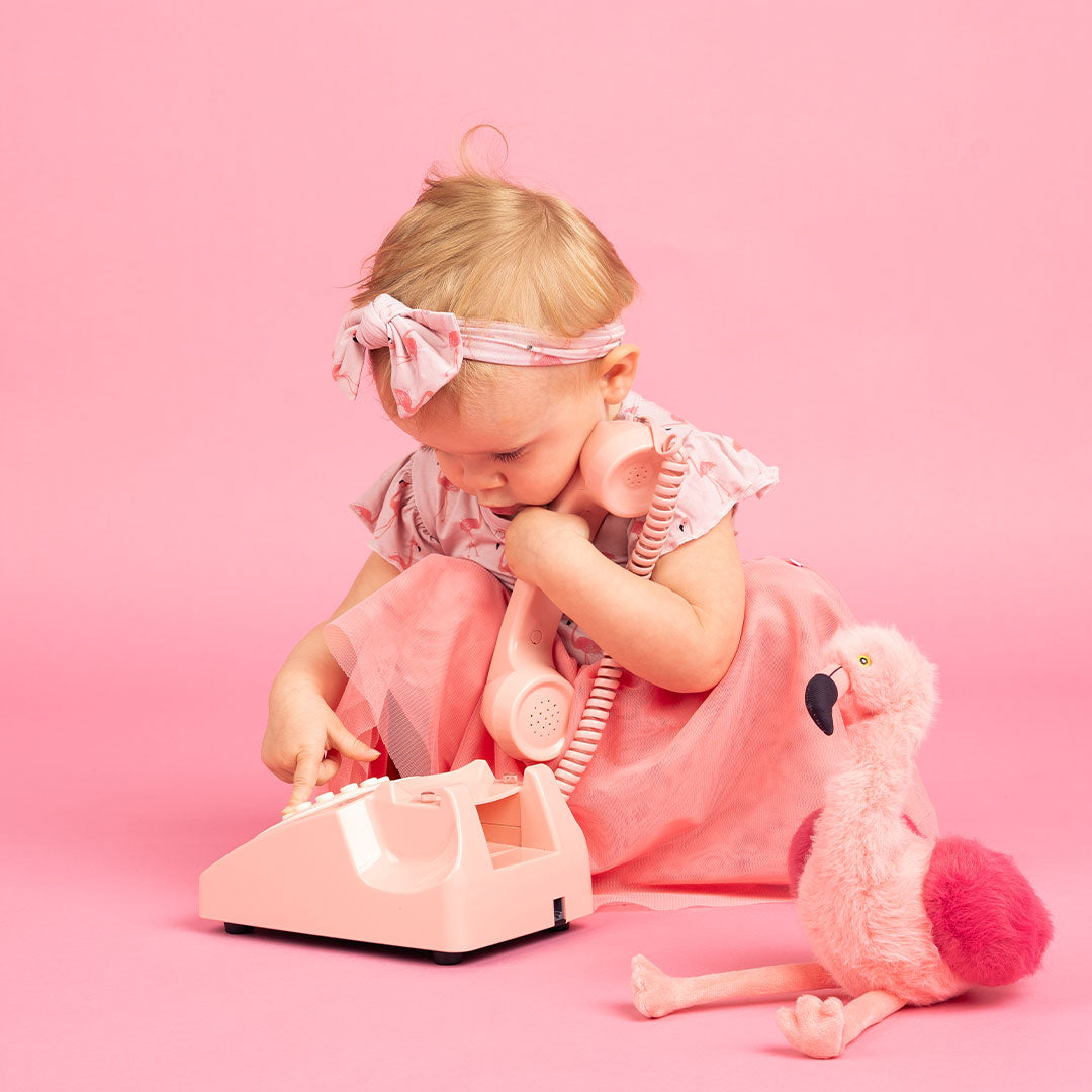 small girl crouched down and plays with an old-fashion pink telephone. she wears the "fancy flamingo" twirl dress and the baby headband. Florence the flamingo sits next to her.