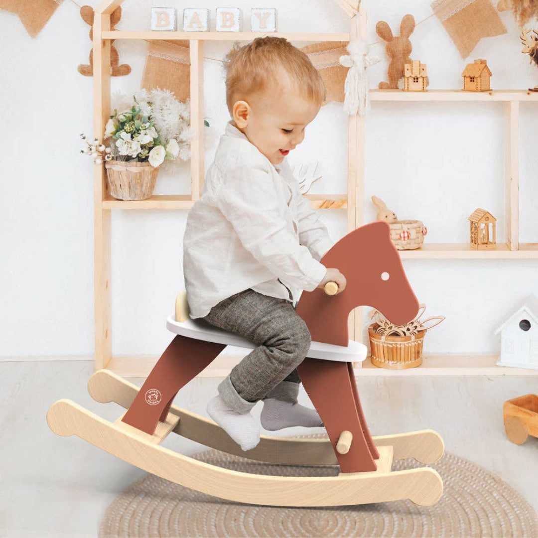 A young child wearing a white shirt, gray pants, and socks is sitting and smiling while riding a wooden rocking horse toy with a rust-orange horse head, white seat, and natural wood base. The background shows a cozy nursery with wooden shelves, soft toys, and decorative items.