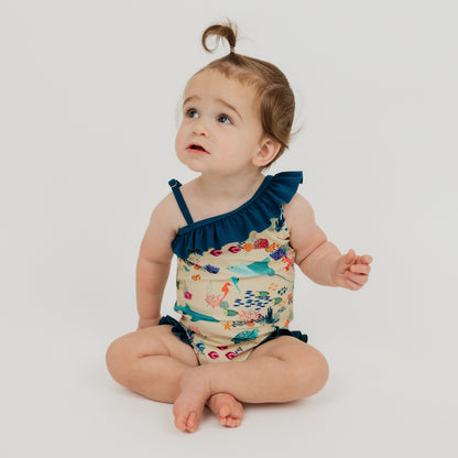 Baby girl sitting down in a white background wearing our Emerson and Friends Coral Reef Ruffle Leg One Piece Girls Swimsuit, featuring a vibrant ocean-inspired print with sharks, sea turtles, and coral, designed with adorable ruffle accents and a secure fit for fun beach and pool days.