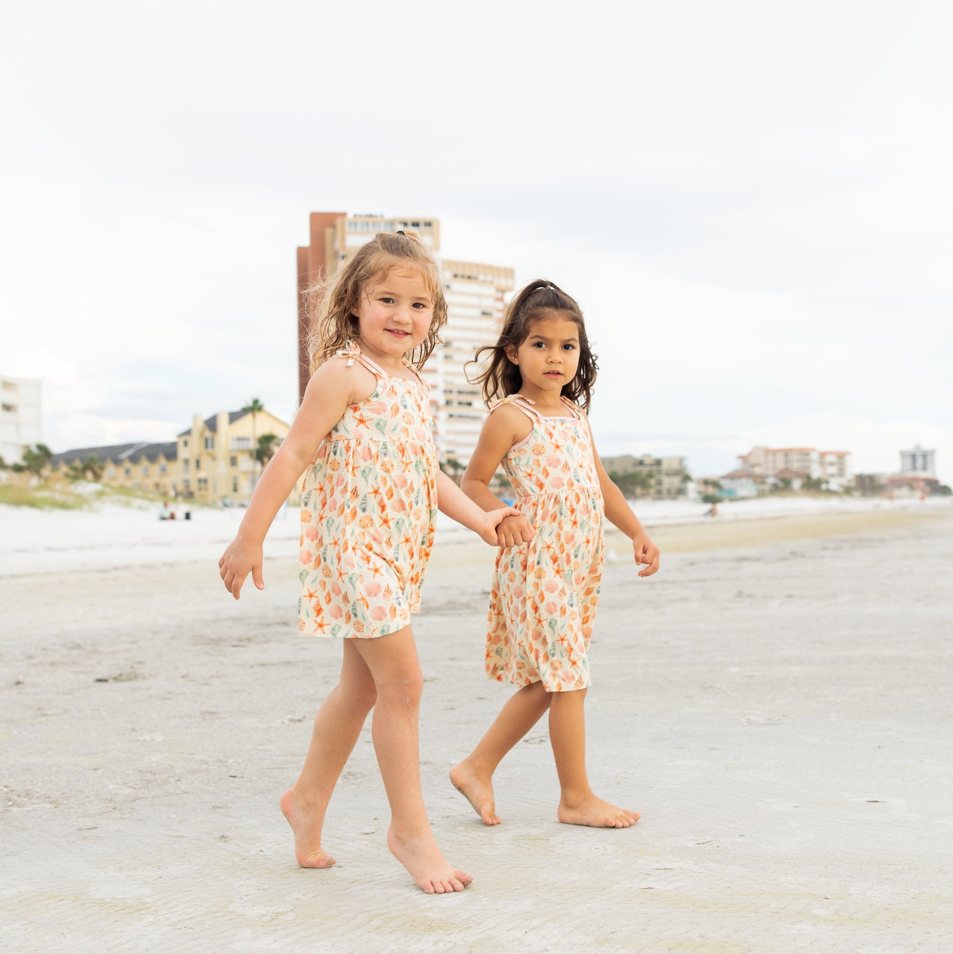 Two little girls holding hands walking on the beach while wearing Sandy Seashell Bamboo Sundresses.