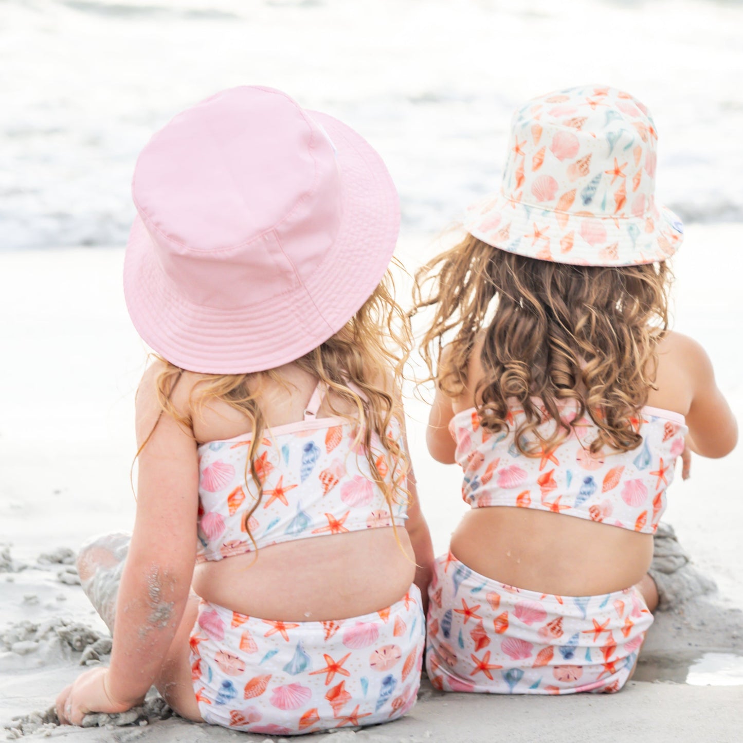 Two girls sitting in the sand wearing the Sandy Seashells Reversible Bucket Hat and Sandy Seashells Bikini Girls Swimsuit. 