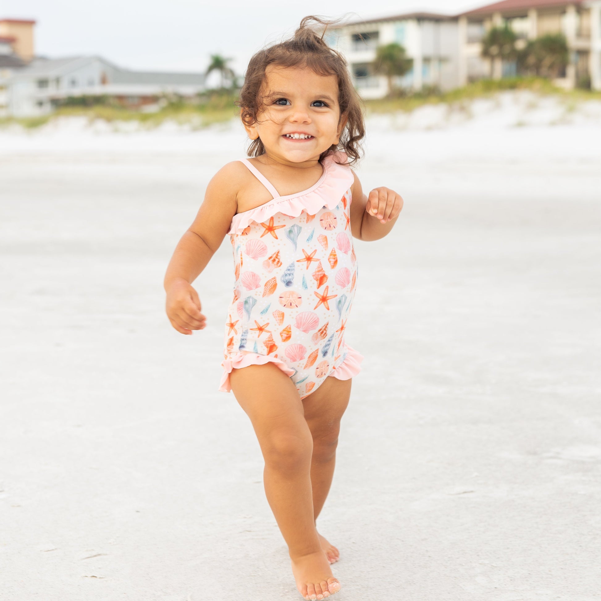 Girl standing in the sand while wearing our  Sandy Seashells Ruffle Leg One Piece Girls Swimsuit. 