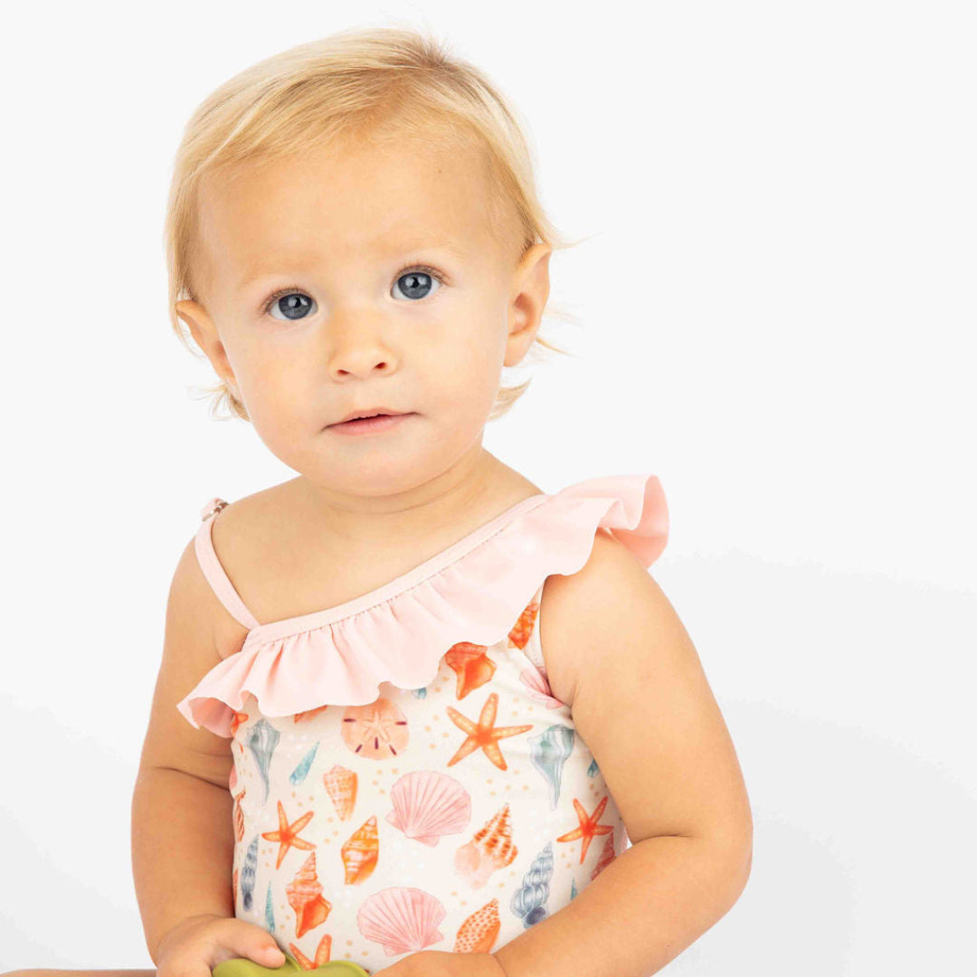 Close up of the baby girl sitting down on a white background wearing our sandy Seashell One piece swim suit with ruffles on the light pink truffles. 